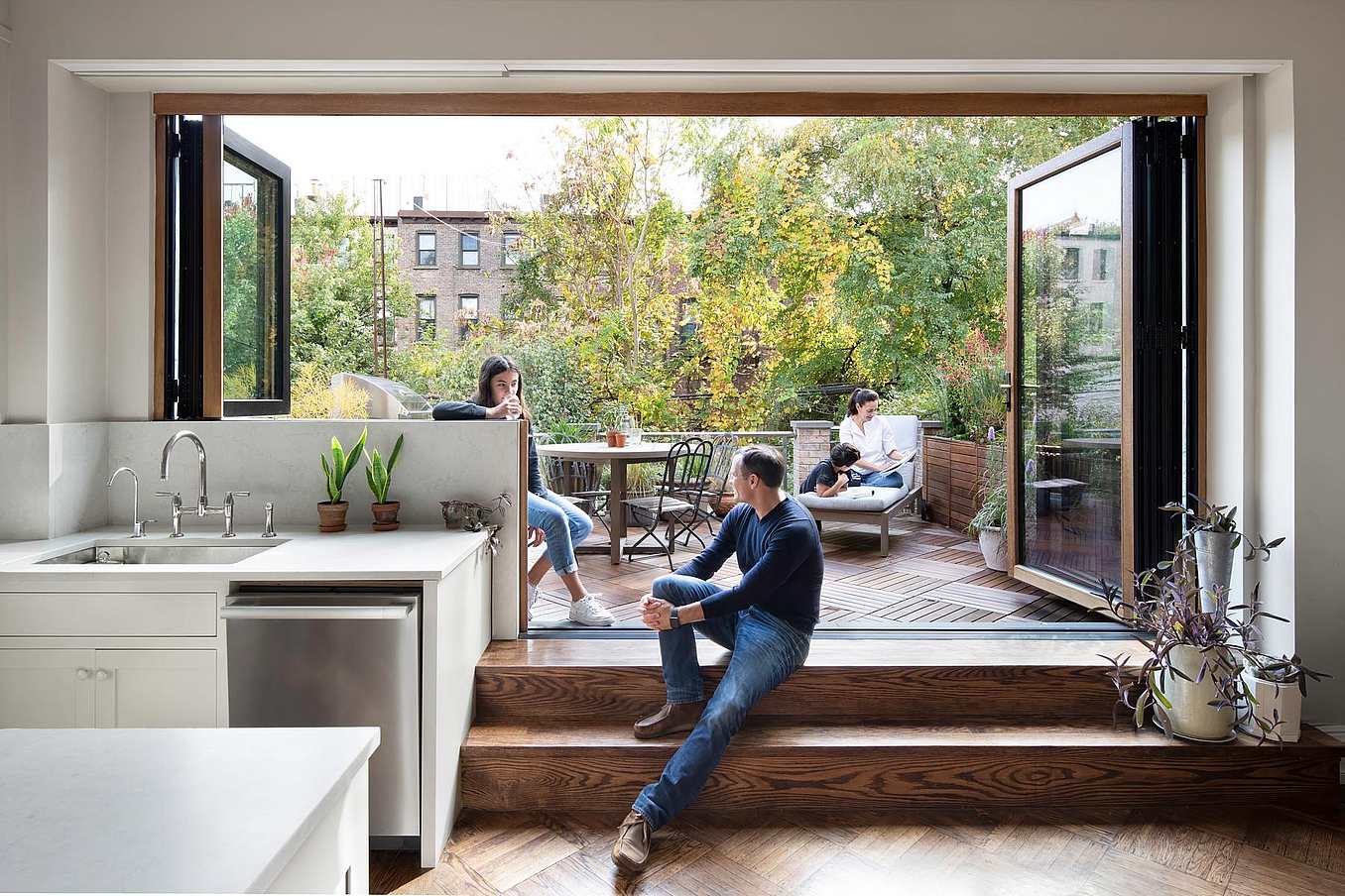 Modern kitchen with retractable glass wall that opens the kitchen to a patio. Three people relax in a modern home with a retractable glass wall that opens the kitchen to a patio; two people sit outside amid greenery, while one sits on indoor steps next to houseplants.