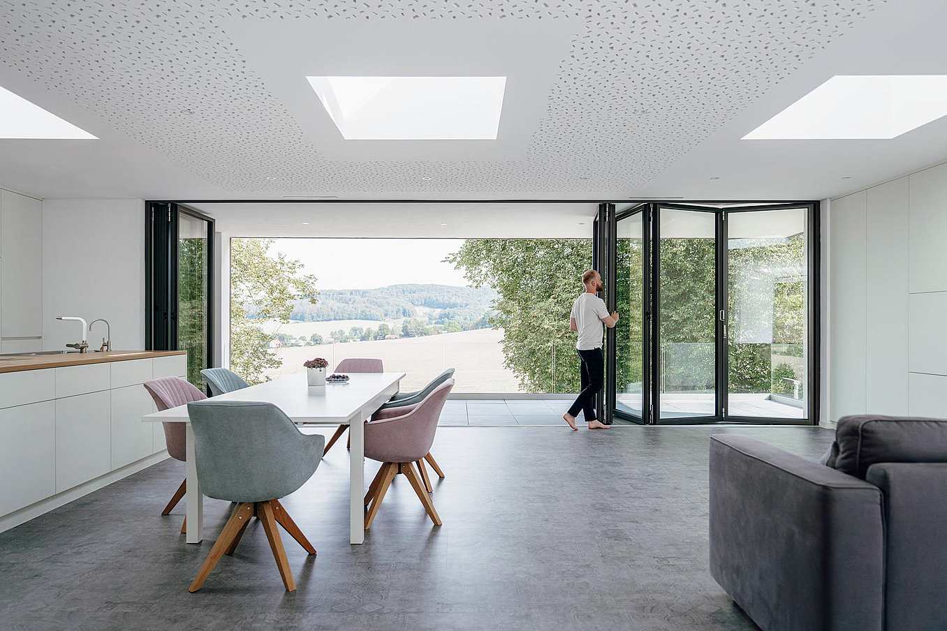 A person opens folding glass doors in a modern, open-plan living and dining area, seamlessly connecting the space to scenic views of trees and fields outside.