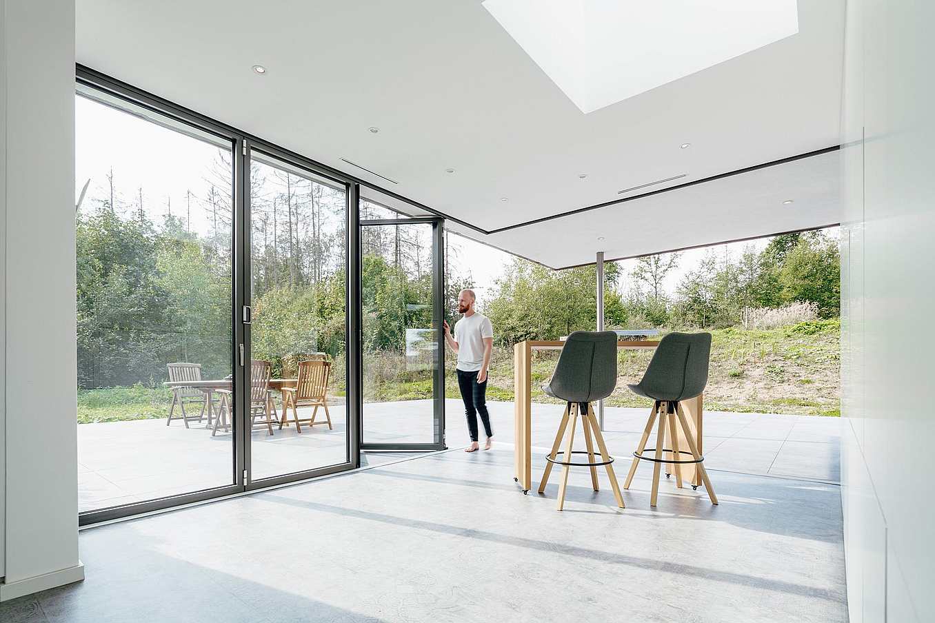 Person standing at open four-panel bifold glass doors, leading from a modern interior to a patio with outdoor furniture and greenery.