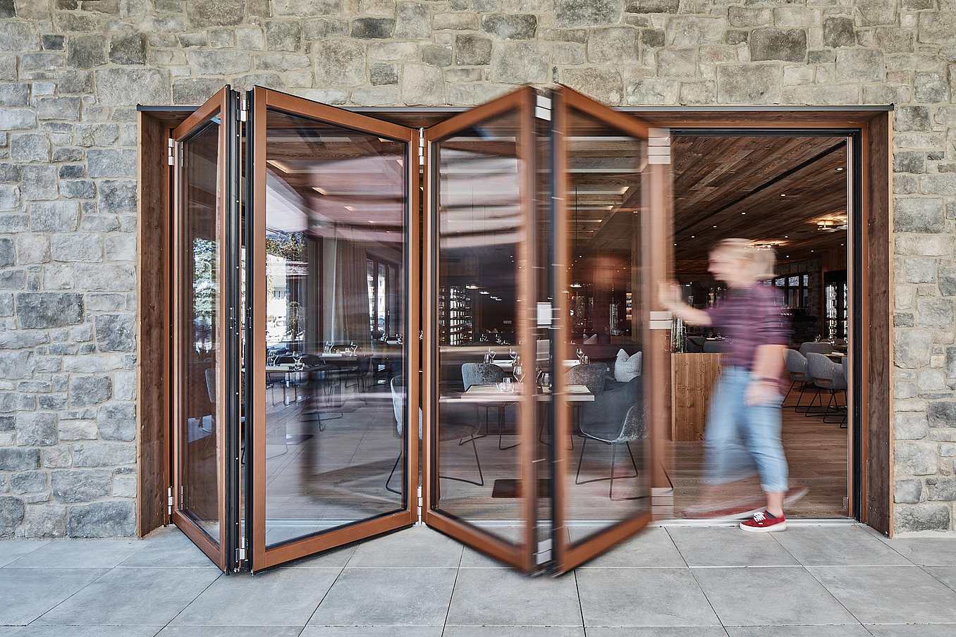 A person opens a glass folding door on a stone building, revealing a modern interior with tables and chairs.