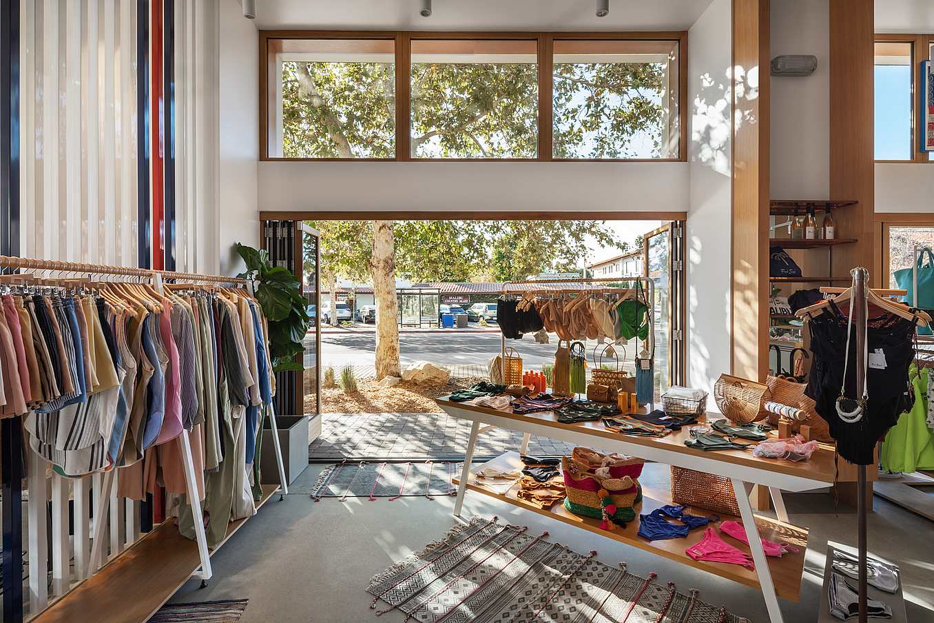 Retail store interior features racks of colorful garments and a central table displaying accessories. Large folding glass doors offer an expansive outdoor view, allowing natural light to stream in, creating a bright and inviting atmosphere.