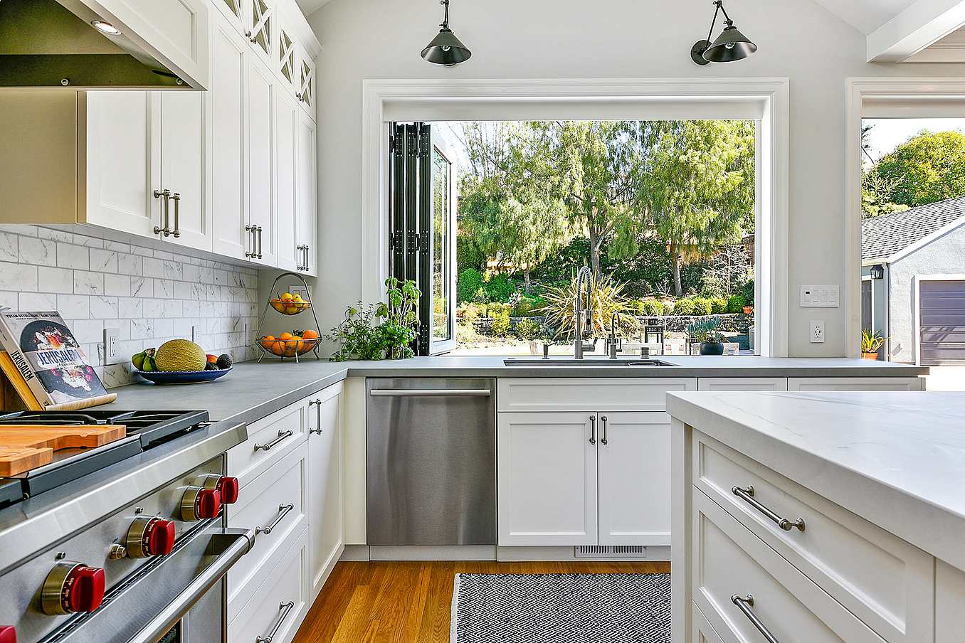 Modern kitchen with white cabinets, stainless steel dishwasher, stove with red knobs, and aluminum clad folding windows overlooking a green backyard.