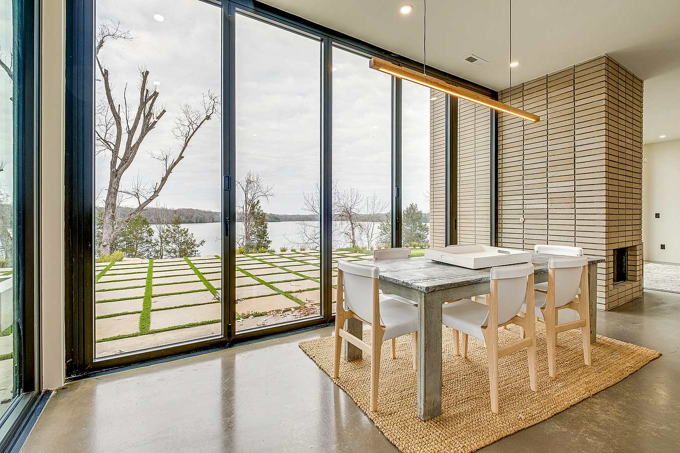 A modern dining area adjacent to floor-to-ceiling glass doors that offer a stunning view of the lake and trees.