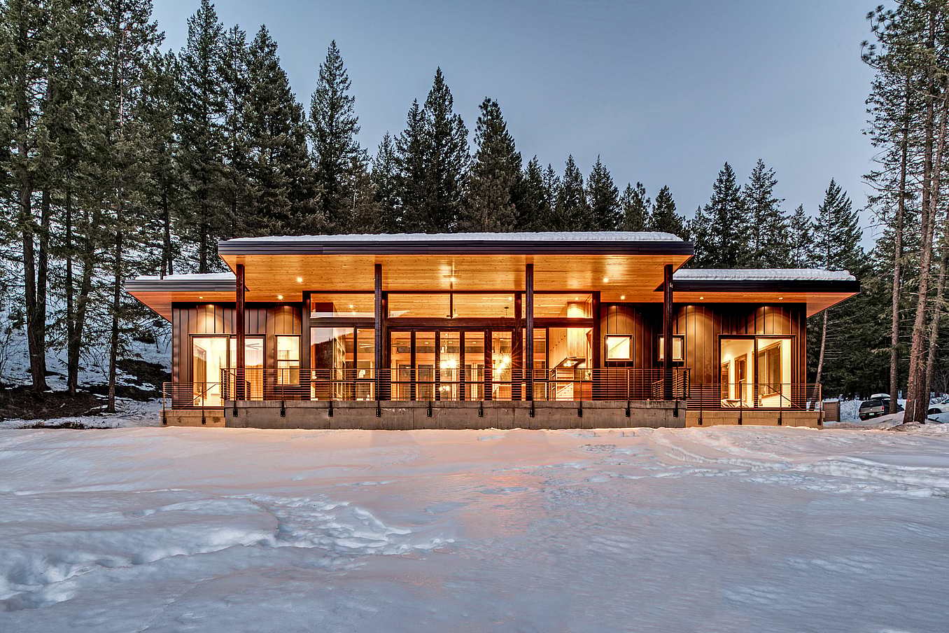 Modern cabin with large windows and warm interior lighting, surrounded by snow and tall pine trees.
