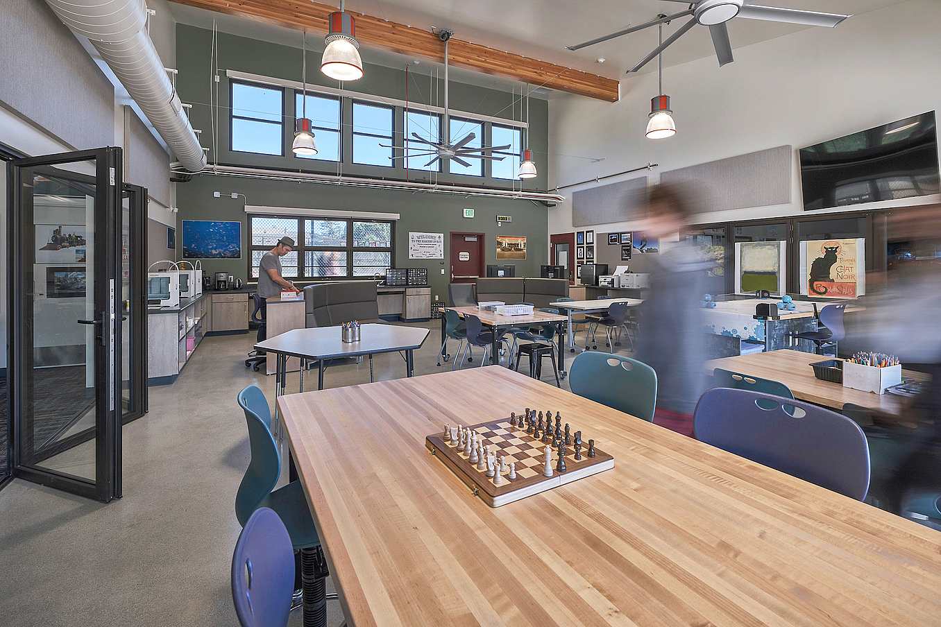 A classroom features tables and chairs, a chessboard on a table, and accordion glass doors leading to another room.