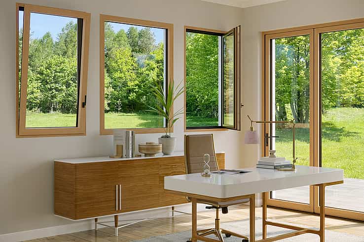 An office with a wooden table features Generation 4 folding glass walls and tilt turn windows, offering a uniform aesthetic and framing views of trees with yellow autumn leaves outside.