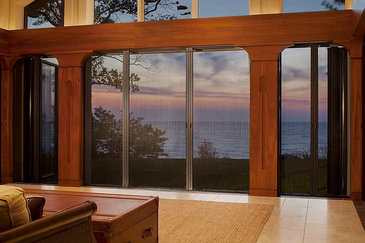 Large glass doors with fully retractable screen panels offer a view of a sunset over a body of water, framed by wooden trim inside a living room.