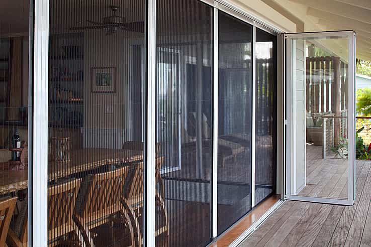 A patio with bifold glass doors and a fully retractable screen door leads from a wooden deck into a dining area with wooden chairs and a table.