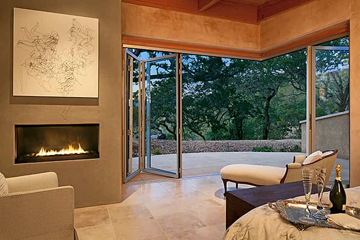 Modern living room with a fireplace, abstract wall art, and open corner accordion glass doors leading to a patio with a view of trees. 