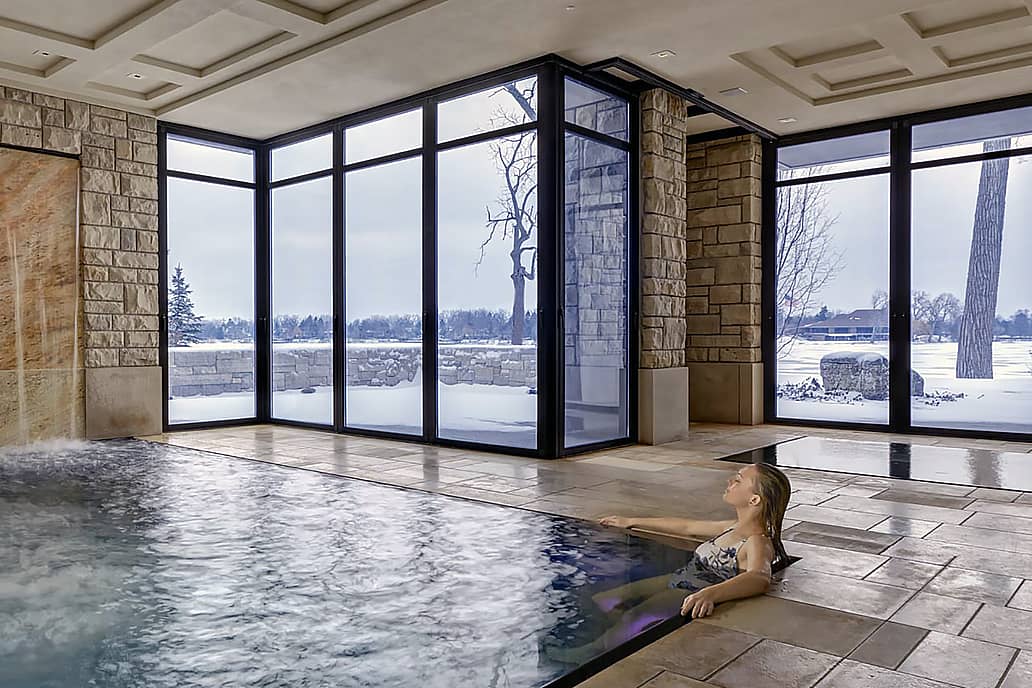 Modern indoor pool with stone waterfall and glass doors overlooking snowy landscape