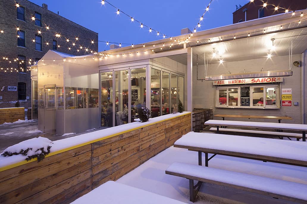 Outdoor restaurant patio with string lights, snow-covered seating, and high-performance glass doors showcasing a cozy indoor restaurant space.