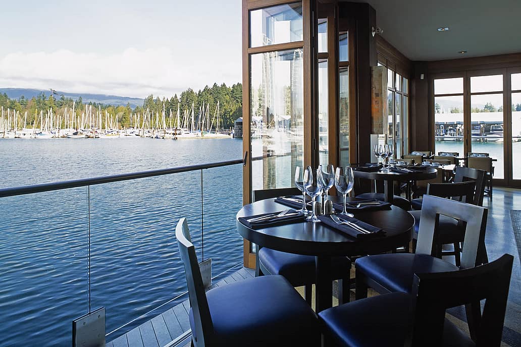 Indoor restaurant seating with set tables and waterfront views of a marina with sailboats, seen through expansive sliding glass doors.