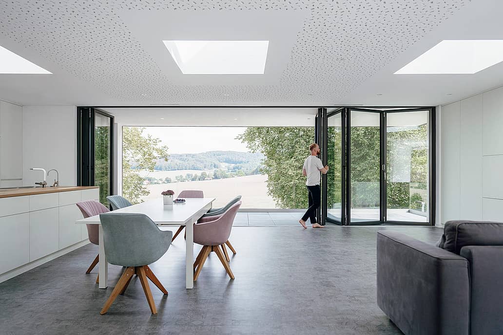 A person opens folding glass doors in a modern, open-plan living and dining area, seamlessly connecting the space to scenic views of trees and fields outside.