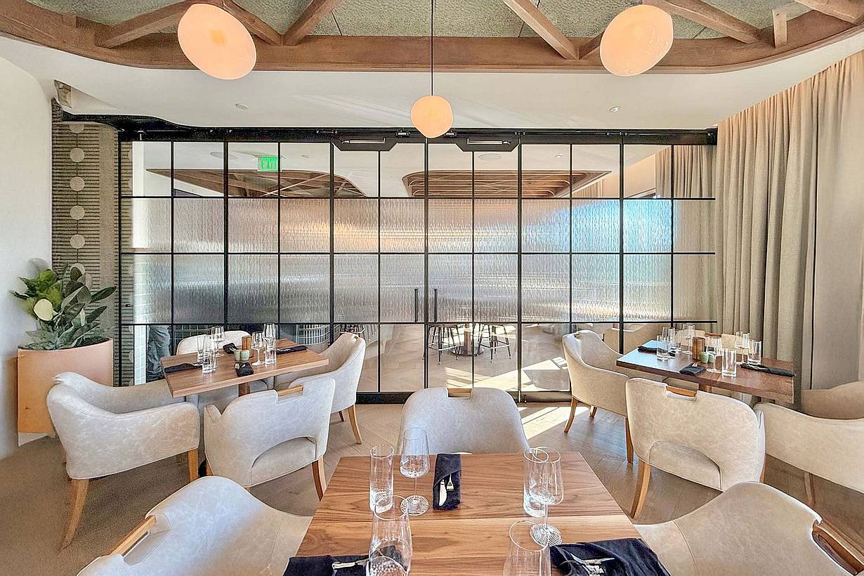 A modern restaurant interior features light wood tables and beige chairs, enhanced by a large glass wall partition for a future amenities area.