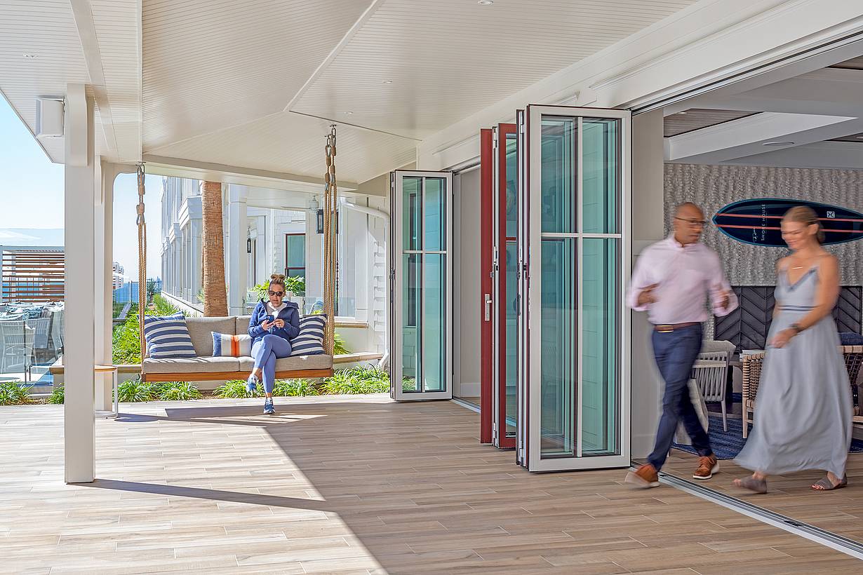 A man sits on a suspended outdoor bench while a man and a woman walk through an open folding glass door into a bright, modern hotel lobby area.