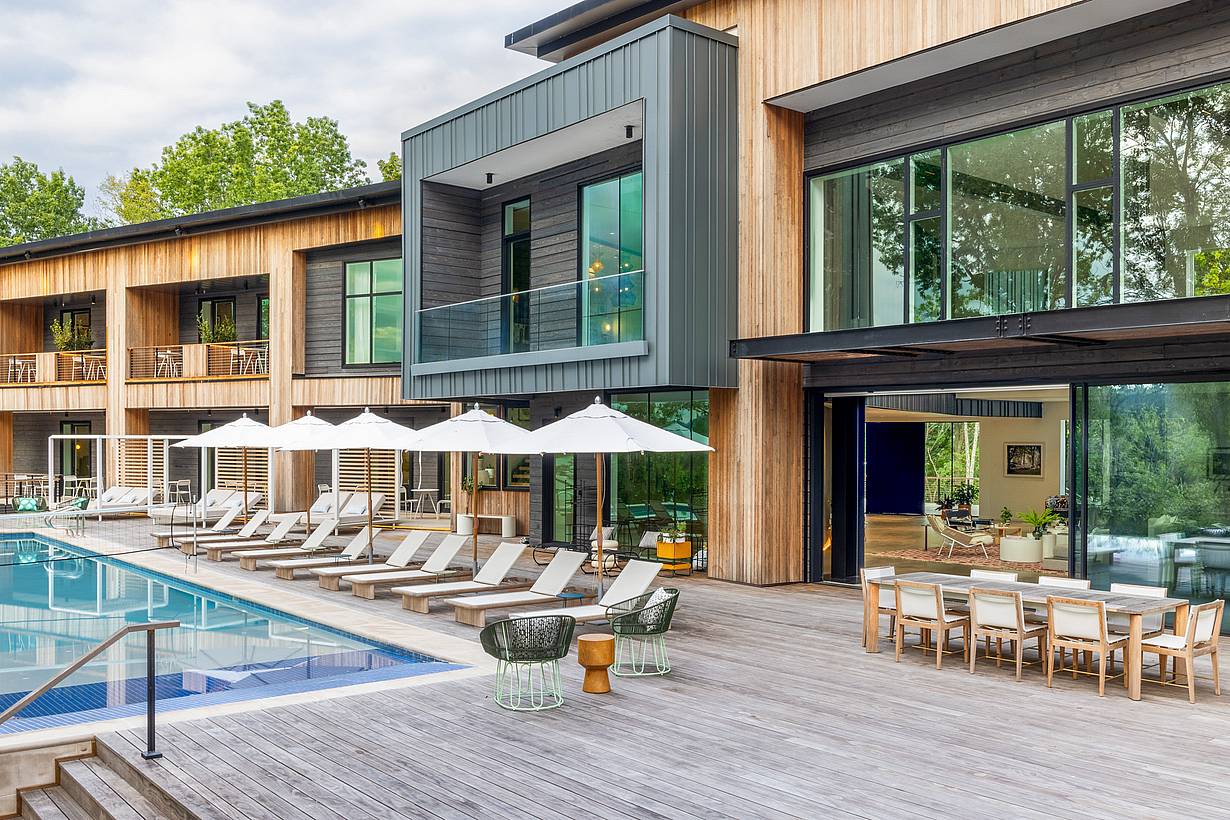 Modern house with large opening glass doors, outdoor swimming pool, lounge chairs with umbrellas, and spacious deck with dining area.