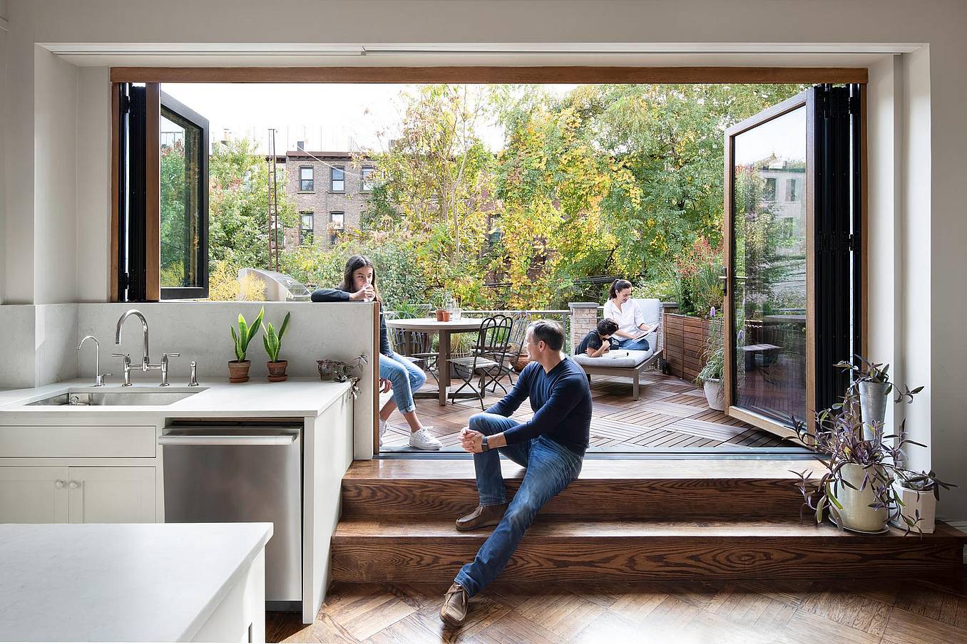 Modern kitchen with retractable glass wall that opens the kitchen to a patio. Three people relax in a modern home with a retractable glass wall that opens the kitchen to a patio; two people sit outside amid greenery, while one sits on indoor steps next to houseplants.