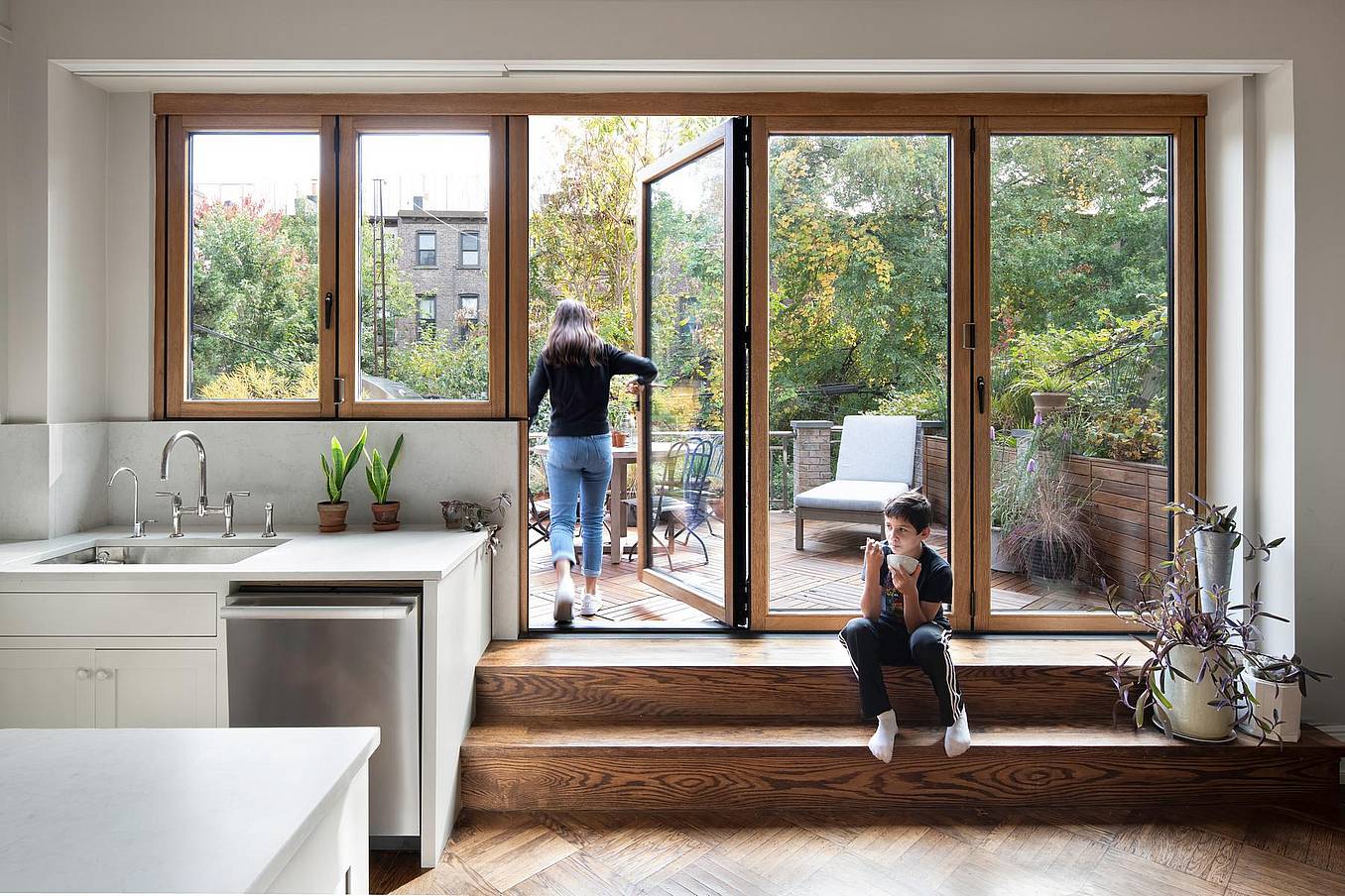 Woman steps onto a patio through wood-framed glass swing doors as a boy sits on wooden steps inside a bright, plant-filled modern kitchen with natural light.
