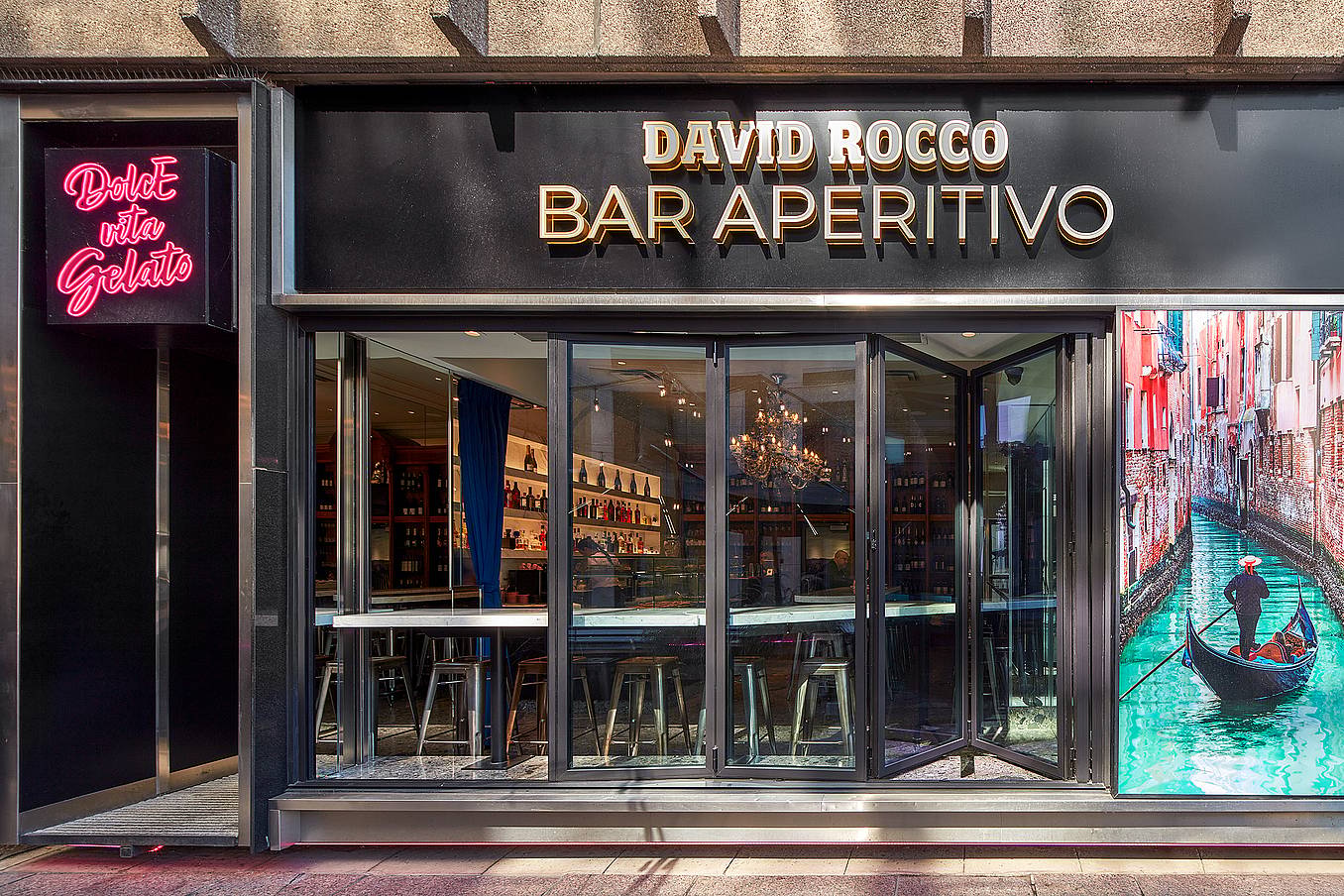 Exterior of 'David Rocco Bar Aperitivo' featuring folding glass doors, a neon 'Dolce Vita Gelato' sign, and a gondola photo, perfect for an authentic Italian experience.