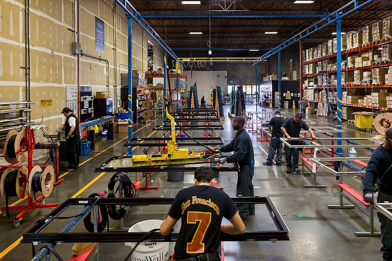 In the NanaWall ISO 9000-certified manufacturing facility in Richmond, CA, workers efficiently assemble products amidst metal tables, hanging tools, and shelves stocked with boxes and materials.
