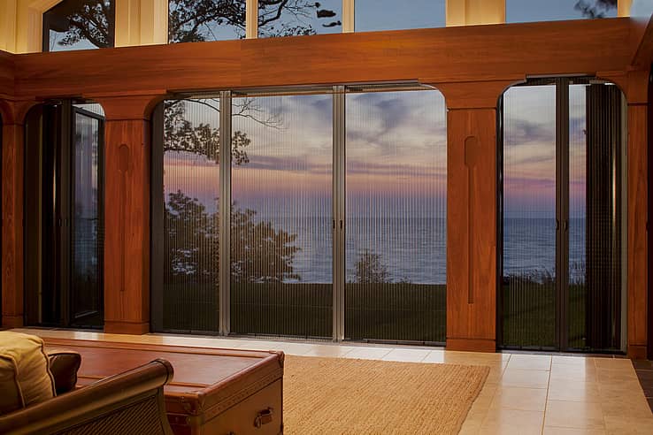 Large glass doors with fully retractable screen panels offer a view of a sunset over a body of water, framed by wooden trim inside a living room.