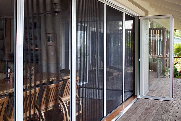 A patio with bifold glass doors and a fully retractable screen door leads from a wooden deck into a dining area with wooden chairs and a table.