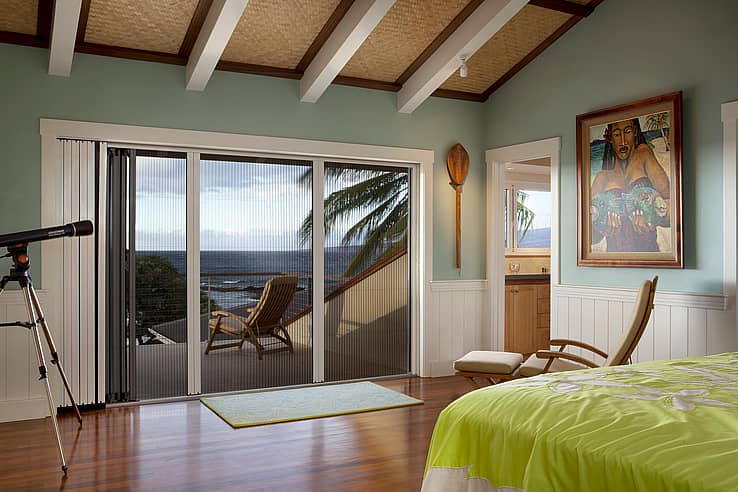 Bedroom with wooden floors, a green bedspread, and folding glass doors with a fully retractable screen opening to a balcony with a lounge chair and ocean view.