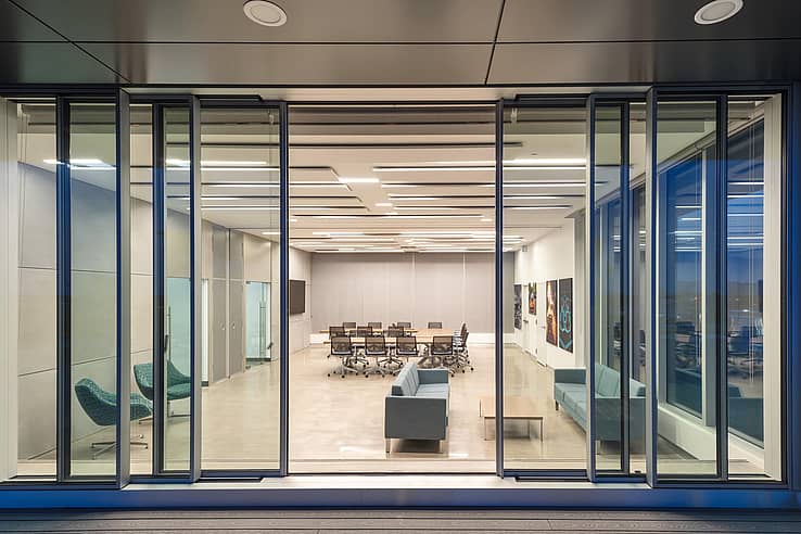 Modern conference room with glass walls, sliding doors, and meeting tables, viewed through large exterior sliding windows.