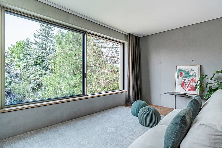Modern living room with large sliding windows, tree views, gray sofa with blue pillows, and framed artwork.
