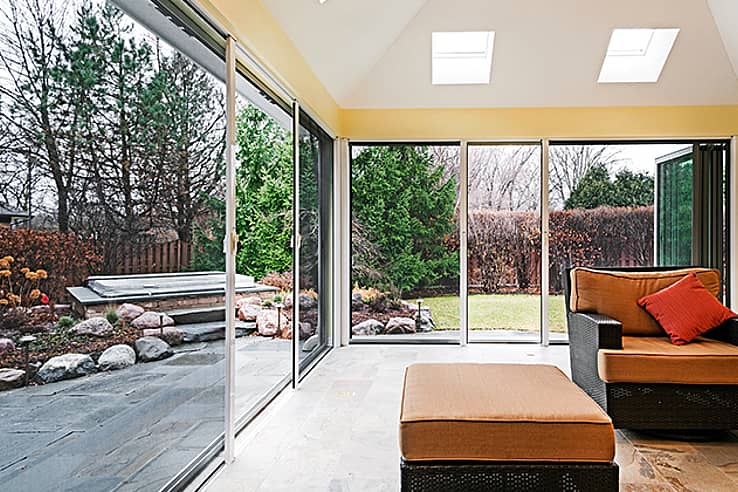 Sunroom with sliding glass doors and screens opens to a landscaped backyard with trees.