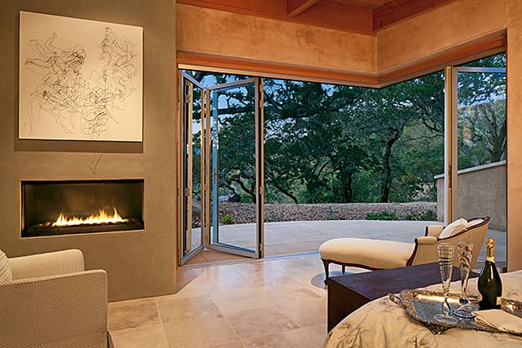 Modern living room with a fireplace, abstract wall art, and open corner accordion glass doors leading to a patio with a view of trees. 