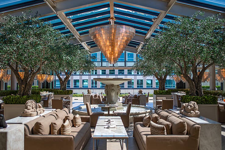 Restoration Hardware rooftop restaurant in Raleigh NC featuring outdoor dining area with sofas and tables around a central fountain under a glass roof with chandeliers and NanaWall SL70 bifold glass doors connecting indoor and outdoor spaces
